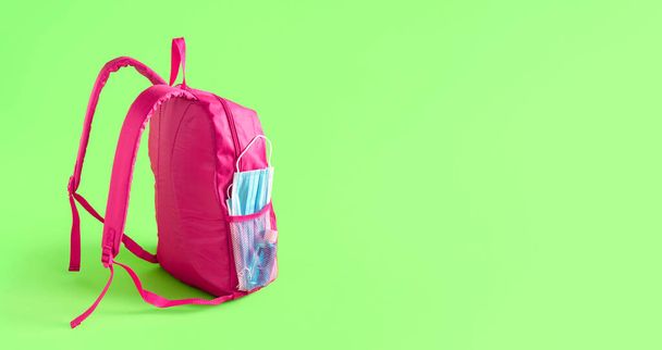 School in pandemic concept. Pink schoolbag with medical masks and hand sanitizer, isolated on green background. New normal in school and travel. - Photo, Image