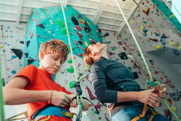 Mother and teenage son at indoor climbing gym near climbing wall. Mother preparing belaying her checking the safety harness boy. Happy parenting concept image.  - Foto, immagini