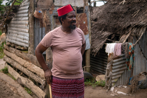 Ein älterer afrikanischer Mann in roter muslimischer Taqiyyah-Fez-Mütze posiert mit einem Stock für Lahme auf einem Hof in der Nähe der einfachen Hütte mit Strohdach in einem kleinen abgelegenen Dorf in Tansania, Insel Pemba, Sansibar - Foto, Bild