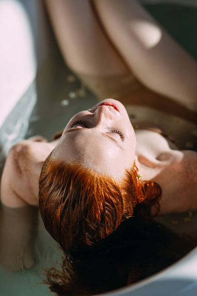 Red-haired young woman lying in the bathtub, gently touching her face by hands - Φωτογραφία, εικόνα