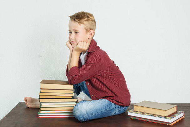 A schoolboy sits on the table near the books, resting his elbows on them and looking away. Education concept - Fotografie, Obrázek