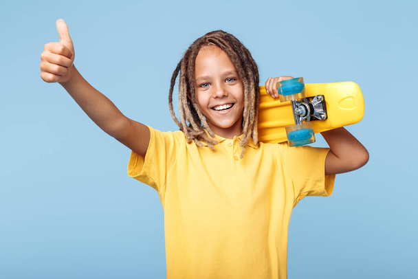 Positive little boy with african dreads holding skateboard on shoulders and showing thumb up over blue background. - Zdjęcie, obraz