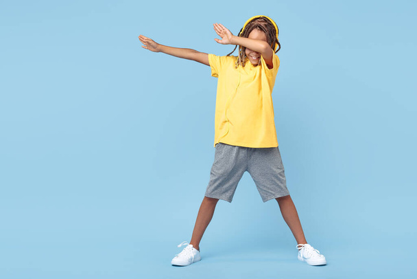 Cheerful little boy with african with headphones dabbing with one leg on soccer ball over blue background. - 写真・画像