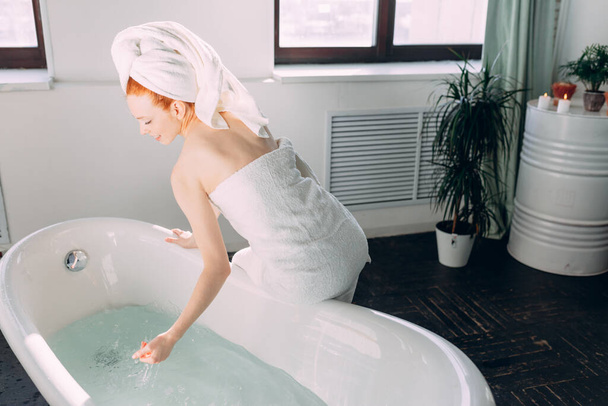 Smiling woman sitting on bathtub edge running the bath, testing water with hand - Photo, Image