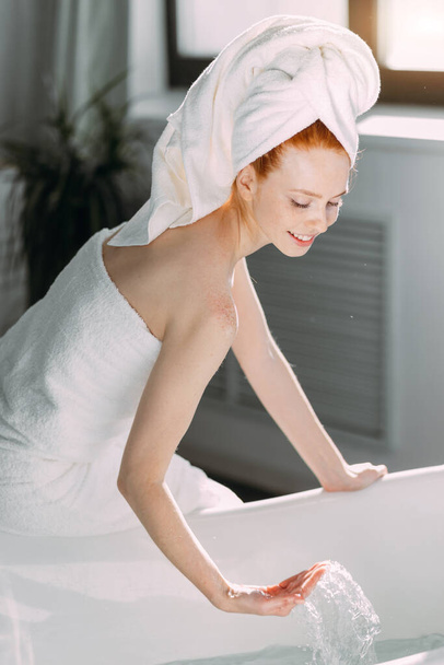 Smiling woman sitting on bathtub edge running the bath, testing water with hand - Valokuva, kuva
