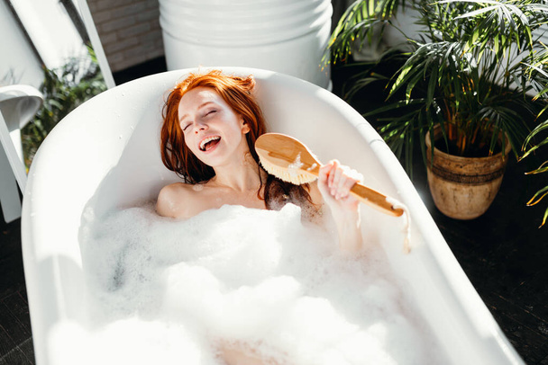 Joyful woman singing using brush as microphone relaxing in bath in bathroom - Fotografie, Obrázek