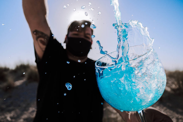 Detailní záběr modrého nápoje ve sklenici a mužského tance v pozadí s hygienickou maskou - koncept nového normálu - Fotografie, Obrázek