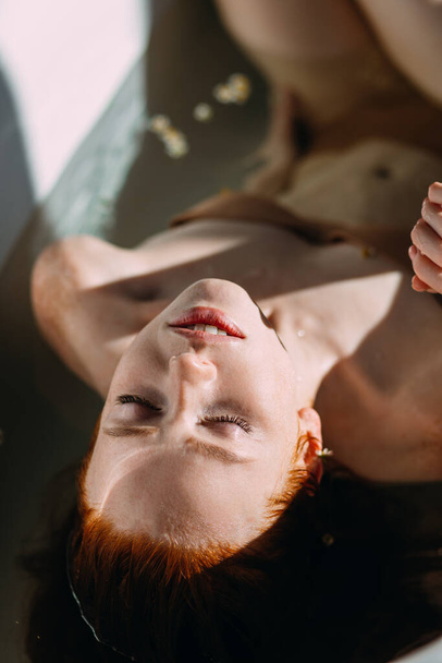 Beautiful seductive young red-haired lady with bare shoulders in bathtub. - Foto, afbeelding