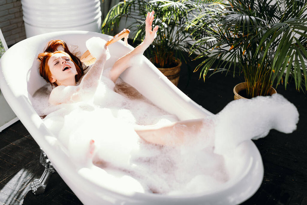 Joyful woman singing using brush as microphone relaxing in bath in bathroom - Valokuva, kuva