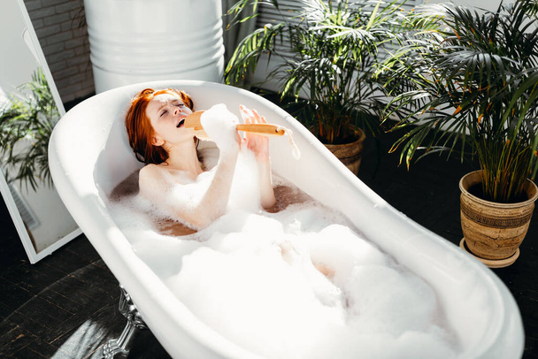 Joyful woman singing using brush as microphone relaxing in bath in bathroom - Foto, immagini