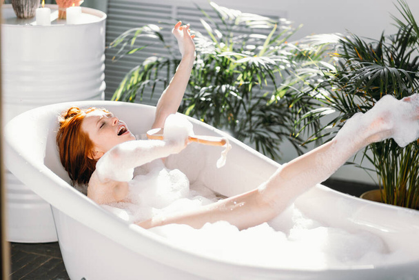 Joyful woman singing using brush as microphone relaxing in bath in bathroom - 写真・画像