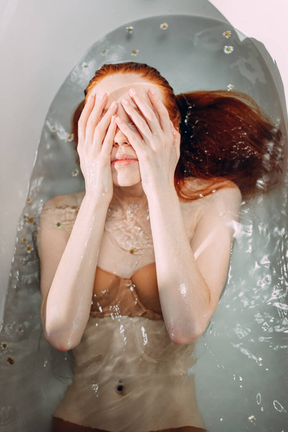 Red-haired young woman lying in the bathtub, gently touching her face by hands - Fotografie, Obrázek