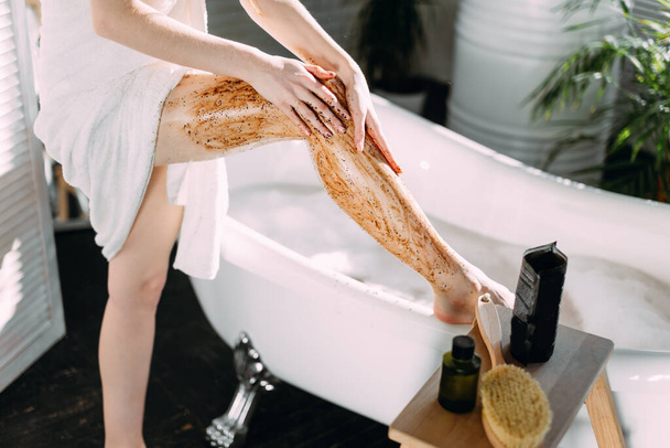 Mujer joven aplicando un exfoliante de café en sus piernas en el baño - Foto, imagen
