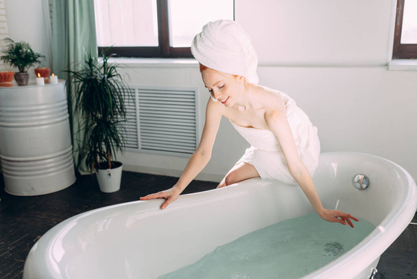 Smiling woman sitting on bathtub edge running the bath, testing water with hand - Foto, Imagen