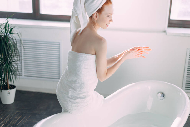 Smiling woman sitting on bathtub edge running the bath, testing water with hand - Foto, imagen