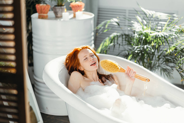 Joyful woman singing using brush as microphone relaxing in bath in bathroom - Fotoğraf, Görsel