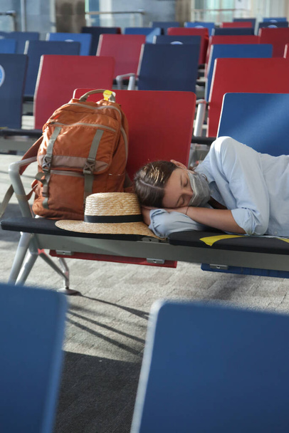 Zmęczona turystka z ochronną maską medyczną śpiąca na bramce terminala czekając na lot. Spóźniony lot, opóźnienie lotu lub odwołanie lotu. Podróżowanie podczas epidemii koronawirusu. - Zdjęcie, obraz