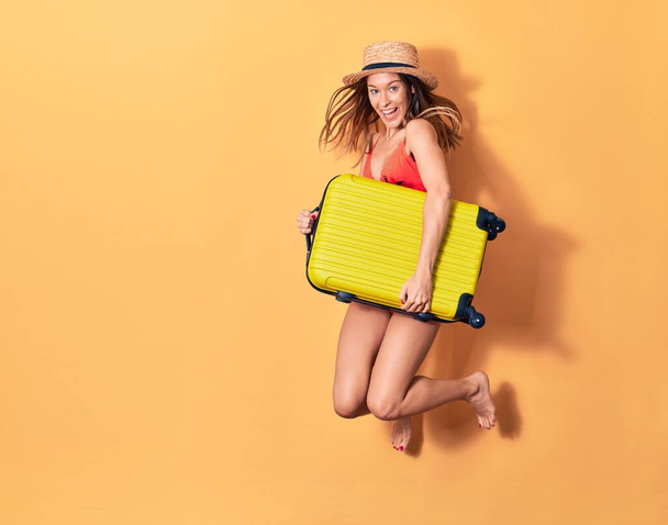 Young beautiful woman on vacation wearing bikini and hat smiling happy. Jumping with smile on face holding cabin bag over isolated background - Foto, afbeelding