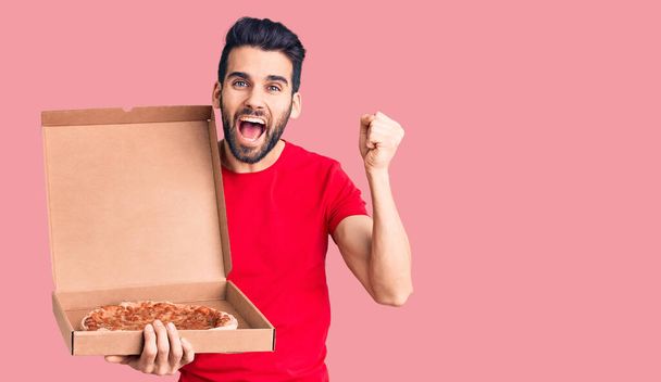 Fiatal, jóképű férfi szakállal, kezében szállítási karton olasz pizza sikoltozó büszke, ünneplő győzelem és a siker nagyon izgatott felemelt karokkal  - Fotó, kép