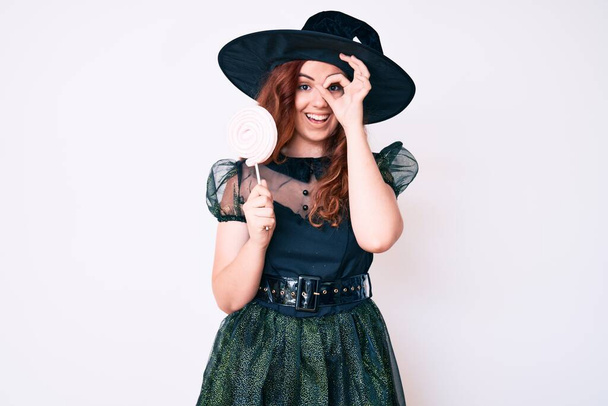 Junge schöne Frau im Hexen-Halloween-Kostüm hält Lutscher lächelnd glücklich tut ok Zeichen mit Hand auf Auge durch Finger schauen  - Foto, Bild