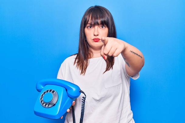Young plus size woman holding vintage telephone pointing with finger to the camera and to you, confident gesture looking serious  - Φωτογραφία, εικόνα