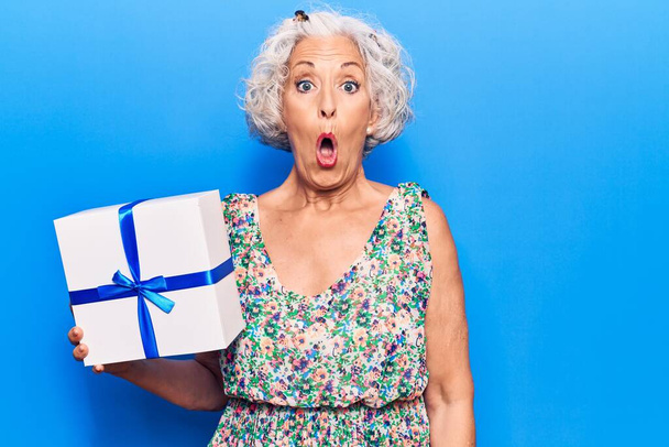 Senior γκρίζα μαλλιά γυναίκα κρατώντας δώρο φοβάται και έκπληκτος με ανοιχτό το στόμα για έκπληξη, δυσπιστία πρόσωπο  - Φωτογραφία, εικόνα