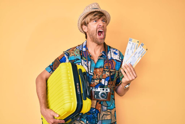 Young blond man wearing summer style holding cabin bag and boarding pass angry and mad screaming frustrated and furious, shouting with anger. rage and aggressive concept.  - Photo, Image