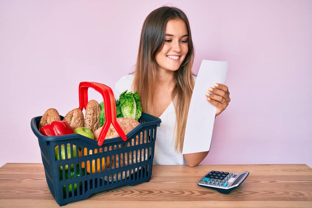Красива біла жінка тримає кошик супермаркетів і продуктовий список посміхається з щасливою і прохолодною посмішкою на обличчі. показ зубів
.  - Фото, зображення