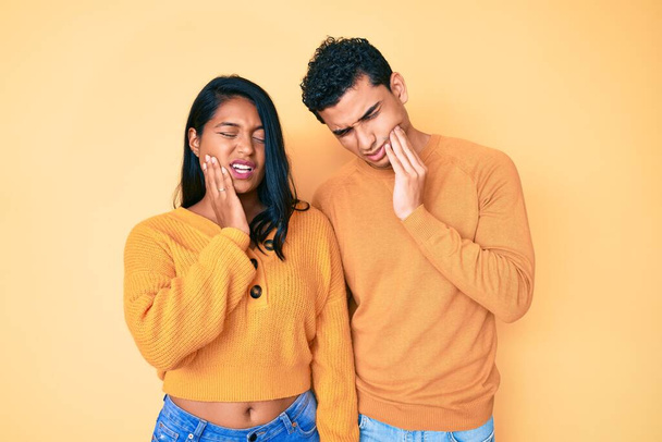 Krásný latinsky mladý pár nosí ležérní oblečení spolu dotýkat ústa s rukou s bolestivým výrazem, protože bolest zubů nebo zubní nemoc na zuby. zubař  - Fotografie, Obrázek