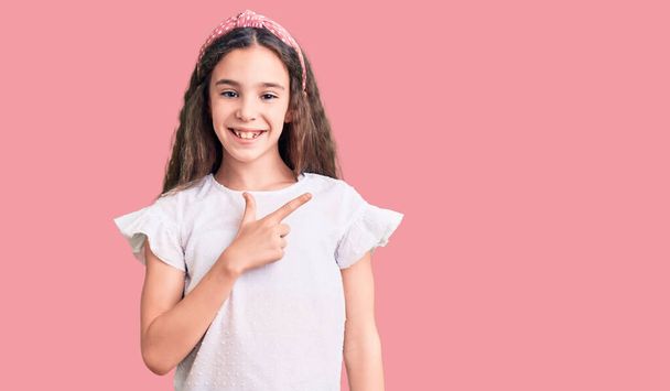 Enfant fille hispanique mignon portant un t-shirt blanc décontracté gai avec un sourire sur le visage pointant avec la main et le doigt sur le côté avec une expression heureuse et naturelle  - Photo, image