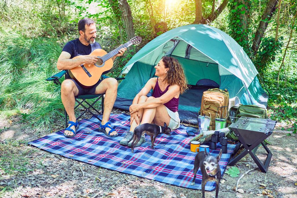 Пара путешественников средних лет, улыбающихся счастливым походом в лесу. Сидя на полу, поет песню, играя на классической гитаре - Фото, изображение