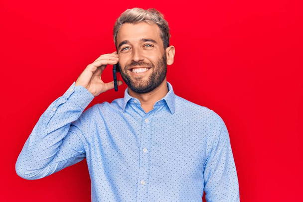 Young handsome blond man with beard having conversation talking on the smartphone looking positive and happy standing and smiling with a confident smile showing teeth - Foto, afbeelding