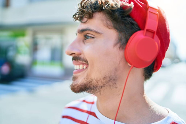Νεαρός καυκάσιος άνδρας χαμογελά χαρούμενος ακούγοντας μουσική χρησιμοποιώντας ακουστικά περπατώντας στην πόλη. - Φωτογραφία, εικόνα