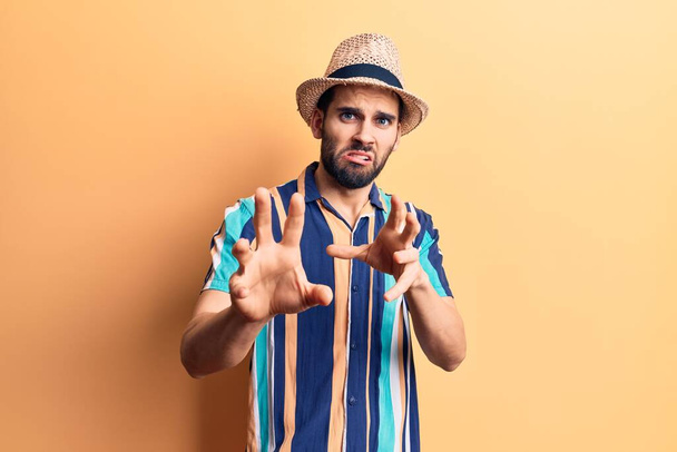 Young handsome man with beard wearing summer hat and shirt disgusted expression, displeased and fearful doing disgust face because aversion reaction. with hands raised  - Photo, Image