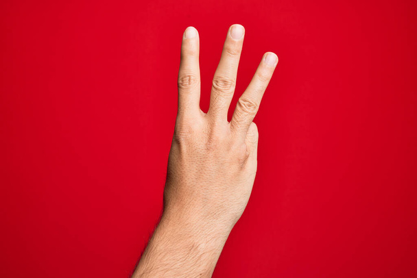 Mano de joven caucásico mostrando dedos sobre fondo rojo aislado contando número 3 mostrando tres dedos - Foto, Imagen