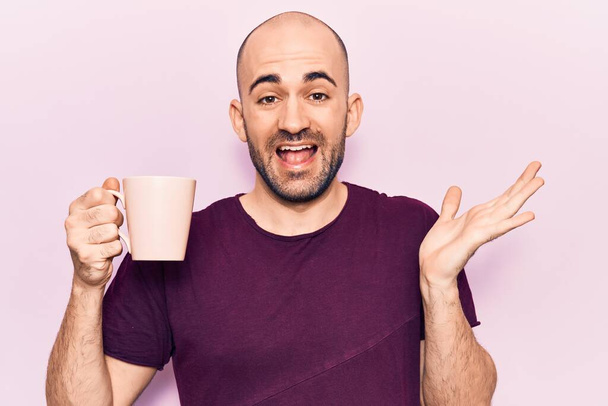 Genç yakışıklı kel adam bir fincan kahve içiyor başarıyı kutluyor mutlu bir gülümsemeyle ve kazanan yüz ifadesiyle elini kaldırarak.  - Fotoğraf, Görsel