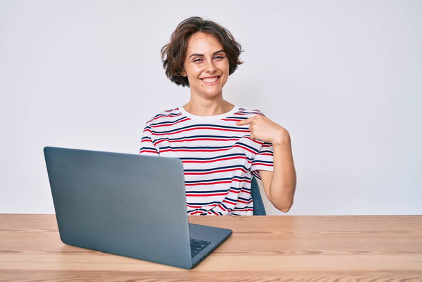 Ofiste çalışan genç İspanyol kadın dizüstü bilgisayarı ile mutlu ve gururlu bir gülümsemeyi işaret ediyor.  - Fotoğraf, Görsel