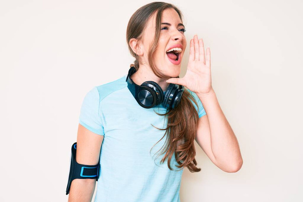 Beautiful young caucasian woman wearing gym clothes and using headphones shouting and screaming loud to side with hand on mouth. communication concept.  - Photo, Image