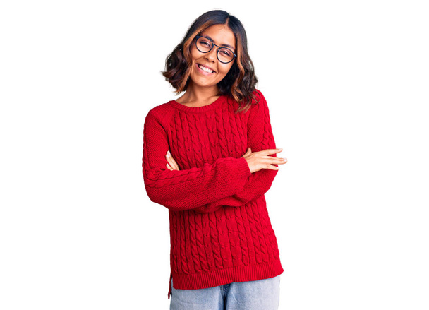 Jonge mooie gemengde ras vrouw draagt rode trui en een bril blij gezicht glimlachend met gekruiste armen kijken naar de camera. positieve persoon.  - Foto, afbeelding