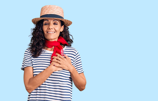 Middle age beautiful woman wearing casual striped t shirt and summer hat smiling with hands on chest with closed eyes and grateful gesture on face. health concept.  - Фото, изображение