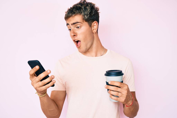 Νεαρός όμορφος άντρας που χρησιμοποιεί smartphone κρατώντας take away καφέ γιορτάζει τρελό και έκπληκτος για την επιτυχία με ανοιχτά μάτια ουρλιάζοντας ενθουσιασμένος.  - Φωτογραφία, εικόνα