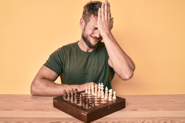 Young caucasian man playing chess sitting on the table surprised with hand on head for mistake, remember error. forgot, bad memory concept.  - Foto, Imagen