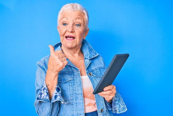 Senior bella donna con gli occhi blu e capelli grigi utilizzando dispositivo touchpad che punta il pollice verso l'alto al lato sorridente felice con la bocca aperta  - Foto, immagini