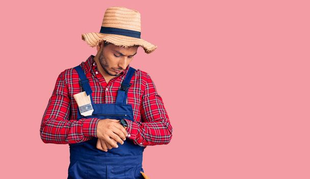 Handsome latin american young man weaing handyman uniform checking the time on wrist watch, relaxed and confident  - 写真・画像