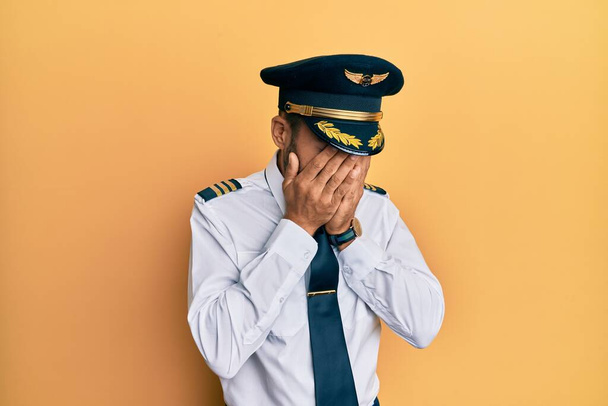 Hombre hispano guapo vistiendo uniforme de piloto de avión con expresión triste cubriendo la cara con las manos mientras llora. concepto de depresión.  - Foto, imagen