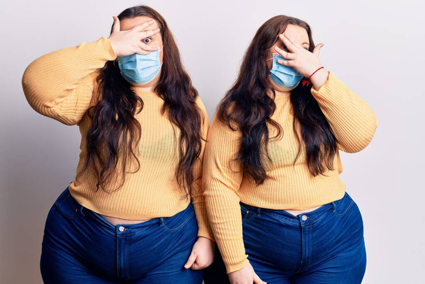 Young plus size twins wearing medical mask peeking in shock covering face and eyes with hand, looking through fingers with embarrassed expression.  - Photo, Image