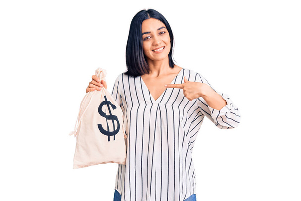 Молодая красивая латиноамериканка держит в руках денежную сумку с символом доллара, указывая пальцем на себя улыбающейся счастливой и гордой  - Фото, изображение