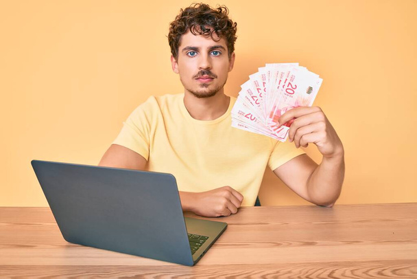 Junger kaukasischer Mann mit lockigem Haar sitzt auf dem Tisch und arbeitet mit Laptop und hält israel-Schekel-Banknoten, denkt Haltung und nüchternen Ausdruck und wirkt selbstbewusst  - Foto, Bild