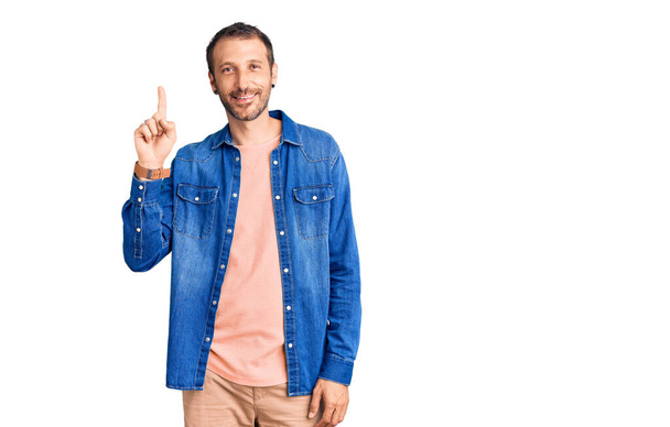 Junger gutaussehender Mann in lässiger Kleidung zeigt mit Finger Nummer eins nach oben und lächelt dabei selbstbewusst und glücklich.  - Foto, Bild