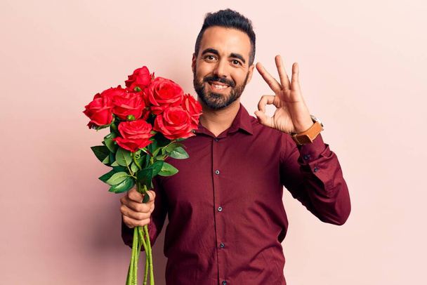 Junger hispanischer Mann hält Blumen in der Hand und winkt mit den Fingern, lächelt freundlich und zeigt ein ausgezeichnetes Symbol  - Foto, Bild